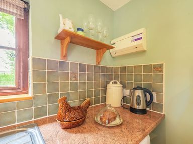 A kitchen corner with tiled walls a wooden shelf holding glassware a ceramic hen container a white canister a kettle and a plate with a covered cake at Cilwen Fach in Cynwyl Elfed
