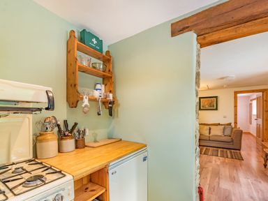 A kitchen countertop with utensils jars and a gas stove next to a fridge with a wooden shelf above and a view into a living room with a sofa at Cilwen Fach in Cynwyl Elfed