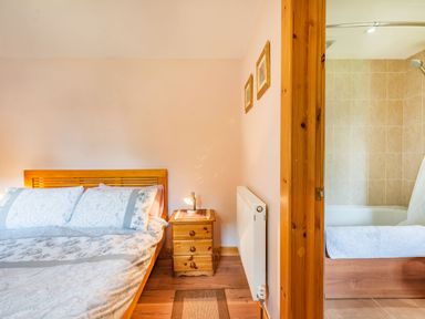 A bedroom with a wooden bed, bedside table, lamp, and a view into a bathroom with a bathtub at Cilwen Fach in Cynwyl Elfed