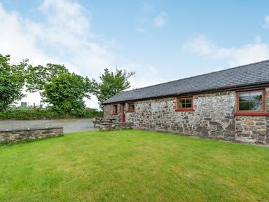 A stone building with three windows a red door a wooden picnic table on a lawn with trees behind at Cilwen Fach in Cynwyl Elfed