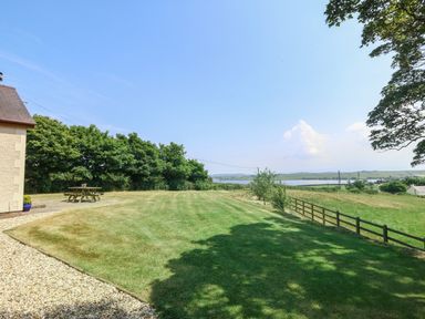 A grassy backyard with a wooden picnic table and trees near a fence overlooking a lake at Fferam Y Llyn in Rhosgoch near Cemaes Bay