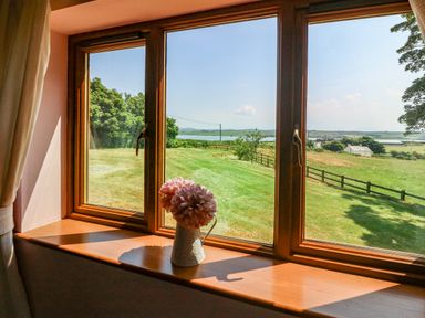 A window with wooden frame looking out onto a grassy field with trees and a small lake a vase with flowers on the windowsill at Fferam Y Llyn in Rhosgoch near Cemaes Bay