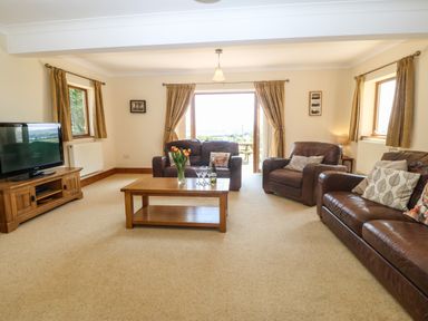 A living room with brown leather sofas a wooden coffee table with flowers and glasses a wooden TV stand with a television and windows with curtains at Fferam Y Llyn in Rhosgoch near Cemaes Bay