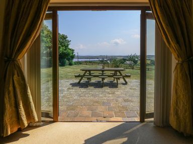 A wooden picnic table on a stone patio outside viewed through open glass doors with curtains at Fferam Y Llyn in Rhosgoch near Cemaes Bay