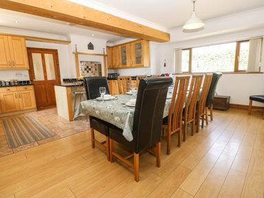 A dining room with a table set for six with wooden and black chairs next to a kitchen with wooden cabinets at Fferam Y Llyn Rhosgoch near Cemaes Bay