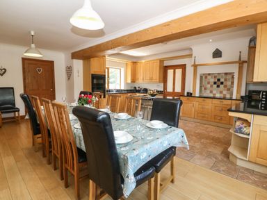 A dining area with a long table set with plates and glasses adjacent to a kitchen with wooden cabinets at Fferam Y Llyn in Rhosgoch near Cemaes Bay