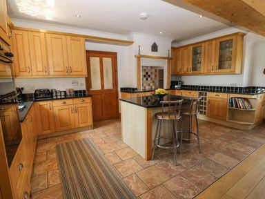 A kitchen with wooden cabinets an island with two metal chairs and a bowl of fruit at Fferam Y Llyn in Rhosgoch near Cemaes Bay