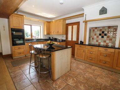 A kitchen with a central island and two bar stools wooden cabinets and tiled floor at Fferam Y Llyn Rhosgoch near Cemaes Bay