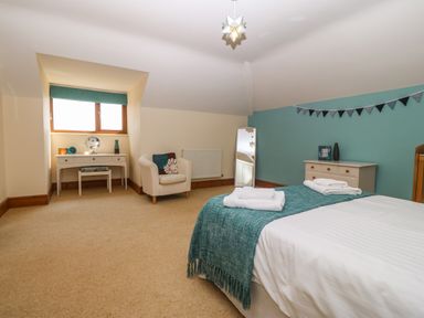 A bedroom with a bed covered in white and teal linens a white armchair a vanity table with a stool a chest of drawers and a mirror at Fferam Y Llyn in Rhosgoch near Cemaes Bay