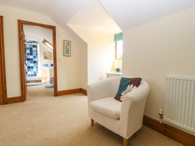 A white armchair with a decorative pillow in a room with beige carpet and a view into a bathroom with blue tiled shower at Fferam Y Llyn in Rhosgoch near Cemaes Bay