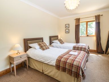 A bedroom with two single beds with plaid blankets and pillows a nightstand with a lamp a window with curtains and two framed pictures on the wall at Fferam Y Llyn in Rhosgoch near Cemaes Bay