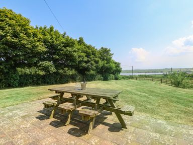 A wooden picnic table with stools on a patio next to a lawn with trees and a view of fields and water at Fferam Y Llyn in Rhosgoch near Cemaes Bay