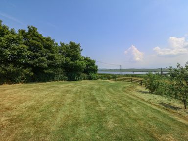A lawn area with trees on the left and a wooden fence overlooking a water body and fields at Fferam Y Llyn in Rhosgoch near Cemaes Bay