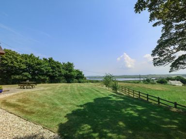 A large lawn with a picnic table trees and a fence overlooking a body of water and fields at Fferam Y Llyn in Rhosgoch near Cemaes Bay