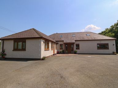A single story house with multiple windows and a tiled roof on a paved driveway at Fferam Y Llyn in Rhosgoch near Cemaes Bay