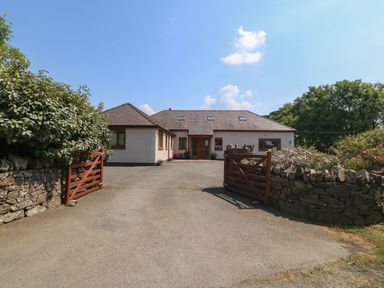A driveway with open wooden gates leading to a large house surrounded by stone walls and bushes at Fferam Y Llyn in Rhosgoch near Cemaes Bay