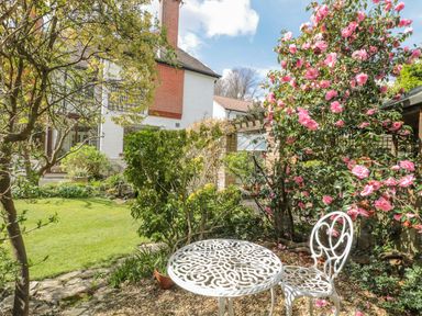 A garden with a white metal table and chair near flowering bushes and a house at West Ridge Boscombe