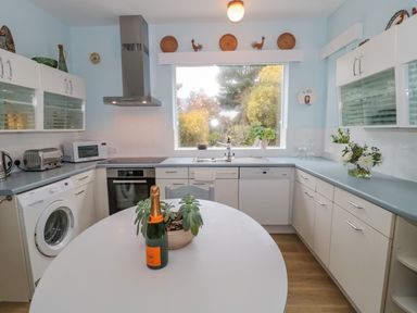 A kitchen with white cabinets a round table with a plant and bottle and a large window showing trees at West Ridge Boscombe