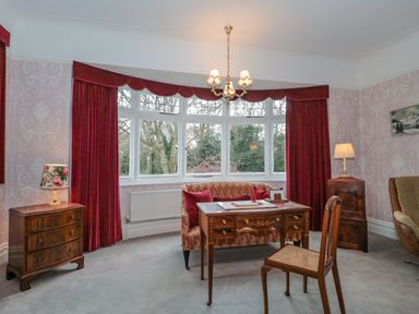 A room with a wooden desk sofa chair two lamps and large bay windows with red curtains at West Ridge in Boscombe