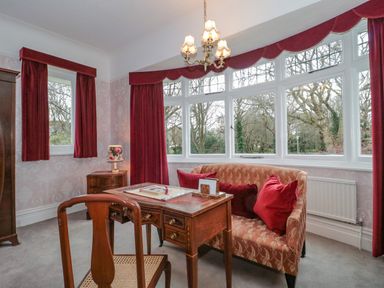 A room with a wooden desk and chair next to a patterned sofa and windows with red curtains at West Ridge in Boscombe