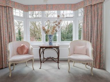 A sitting area with two chairs a small wooden table with a vase with dried flowers in front of a large bay window with patterned curtains at West Ridge in Boscombe