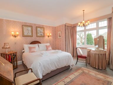A bedroom with a bed with white linens and pink pillows two framed pictures above the bed a vanity with a mirror near a window and a chair at West Ridge in Boscombe