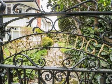 A black wrought iron gate with gold lettering and a stone path leading to a house surrounded by plants and bushes at West Ridge in Boscombe