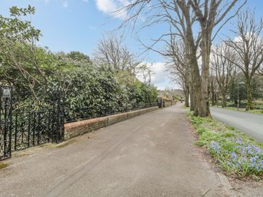 A sidewalk next to a road with trees and flowers along the edge and a black iron gate with bushes behind it at West Ridge in Boscombe