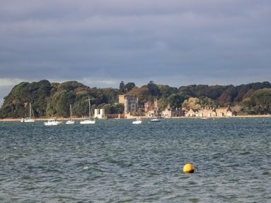 A body of water with boats anchored near a wooded shore with buildings and a castle in the background at West Ridge in Boscombe