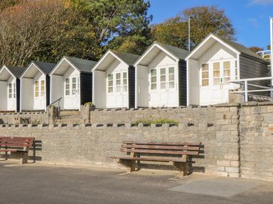 A row of small beach huts with white doors on a stone terrace with two wooden benches in front at West Ridge in Boscombe