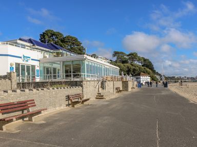 A cafe with glass windows beside a paved walkway and benches near a beach with people walking at West Ridge in Boscombe