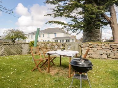 An outdoor garden area with a table covered by a cloth and wooden chairs near a barbecue grill at Tremanners in St Agnes