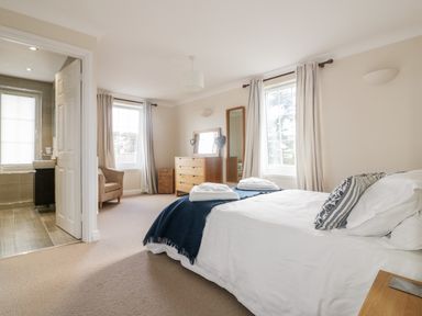 A bedroom with a bed, two windows with curtains, a chest of drawers, a mirror, an armchair, and an open door to a bathroom at Tremanners in St Agnes