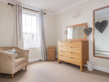 A room with a beige armchair, a wooden dresser with a mirror, a wicker basket, and striped curtains at Tremanners in St Agnes