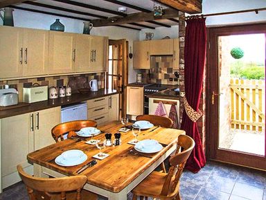 A kitchen with wooden dining table set with plates and glasses and cream cabinets with cooking appliances at Owl Cottage in Napton on the Hill near Southam