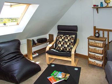 A room with a black leather chair a wooden armchair with a checkered cushion a small wooden cabinet with woven drawers a low black table with books and a skylight window at Owl Cottage in Napton-on-the-Hill near Southam