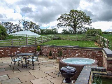 An outdoor patio with a round table and four chairs under a white umbrella a hot tub a small grill potted plants and a grassy area with wooden fencing at Owl Cottage in Napton-on-the-Hill near Southam