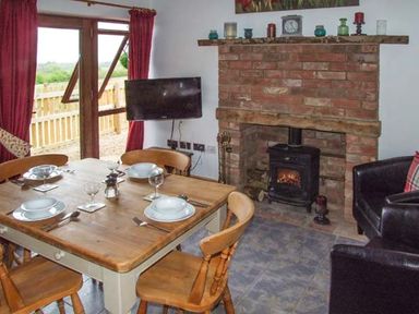 A dining area with a wooden table set for four and a brick fireplace with a stove and a wall-mounted TV at Owl Cottage in Napton-on-the-Hill near Southam