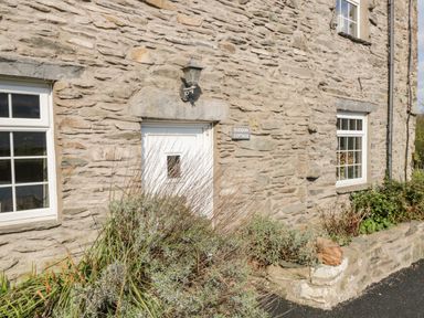 Stone exterior wall of Duddon Cottage with white door and windows surrounded by shrubs and plants at Broughton-in-Furness