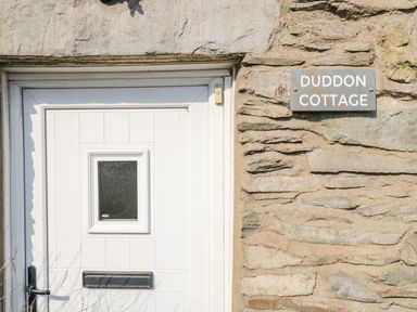 A white door with a small square window and a mailbox on a stone wall with a sign that reads Duddon Cottage at Broughton-in-Furness