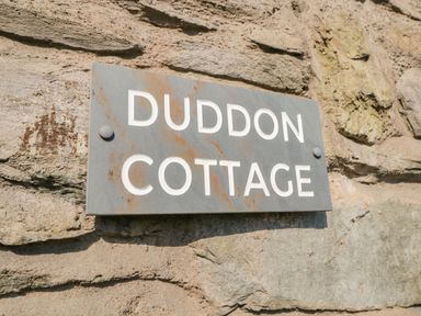A close up of a gray metal sign with white letters saying Duddon Cottage mounted on a stone wall at Duddon Cottage in Broughton-in-Furness
