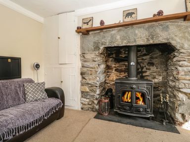 A room with a stone fireplace and wood burning stove next to a sofa with a patterned cushion at Duddon Cottage in Broughton-in-Furness