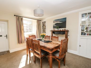A dining room with a wooden table and six chairs a window with striped curtains a door a cupboard with glass doors and books a mantel with a plant and a framed picture at Duddon Cottage in Broughton-in-Furness