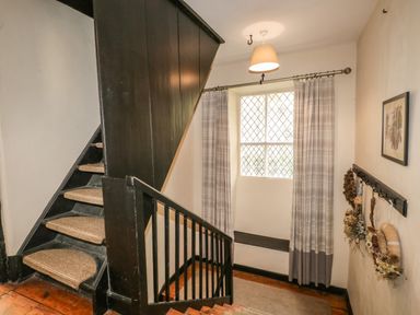 A staircase with black banisters and brown carpet rods near a window with checkered curtains and a wall with hanging decorations at Duddon Cottage in Broughton-in-Furness