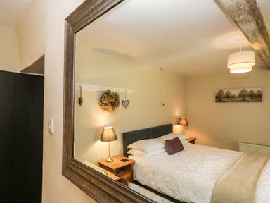 A bedroom with a bed lamps a wooden side table a framed picture and a coat rack reflected in a large wall mirror at Duddon Cottage in Broughton-in-Furness