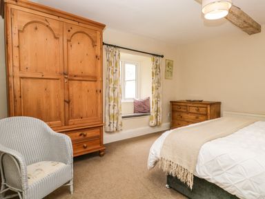 A bedroom with a wooden wardrobe a chest of drawers a window with curtains and a chair at Duddon Cottage in Broughton-in-Furness