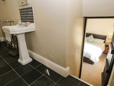 A bathroom with a white pedestal sink and black tiled floor next to a doorway leading to a bedroom with a bed and bedside table at Duddon Cottage in Broughton-in-Furness