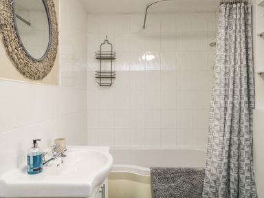 A bathroom with a white sink, a round mirror, a bathtub with a shower curtain and a wall-mounted wire rack at Duddon Cottage in Broughton-in-Furness