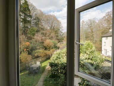 A view through an open window showing a garden with outdoor table and chairs and trees at Duddon Cottage in Broughton-in-Furness