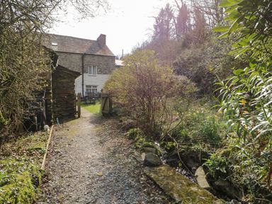 A gravel pathway leading to a stone cottage with garden furniture and surrounding trees and bushes at Duddon Cottage in Broughton-in-Furness